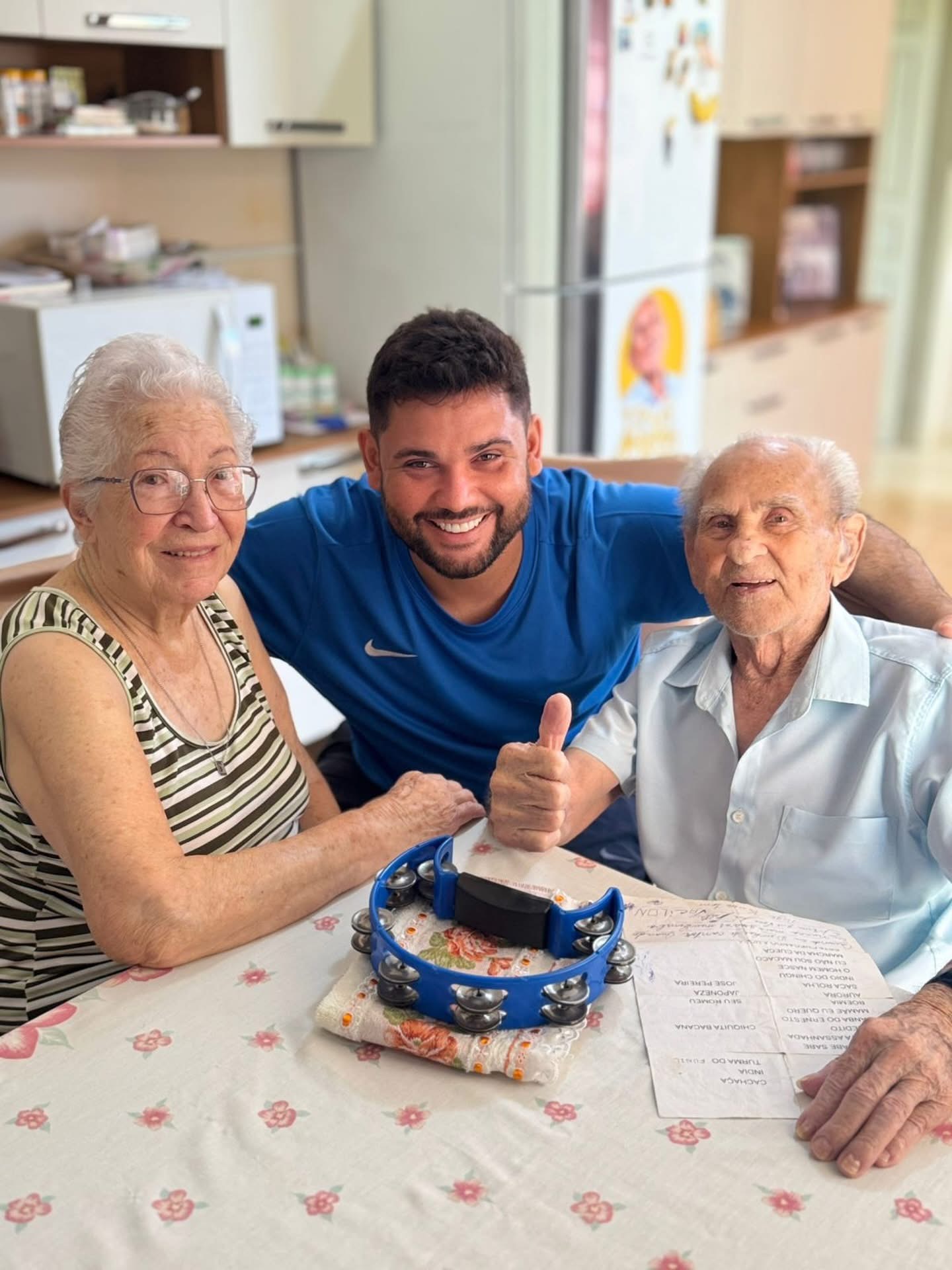 Caio entre a Vovó Elza e o Vovô Anésio, com o pandeiro sobre a mesa